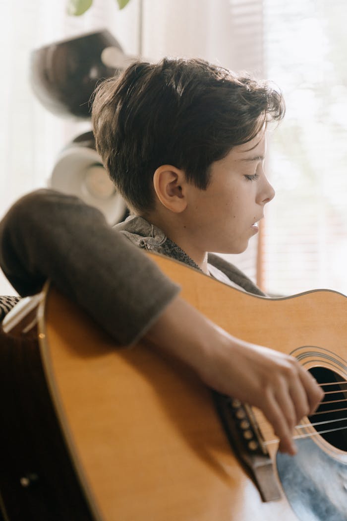 A boy plays an acoustic guitar indoors, focused and intent in natural light.