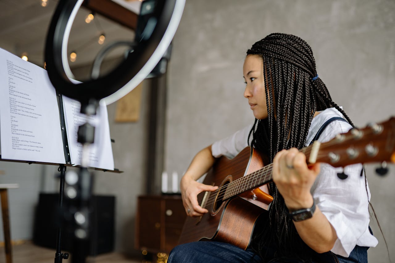 Young woman playing acoustic guitar, reading music sheets indoors with ring light